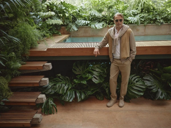Mature adult male model poses in a tan jacket and pants near a modern, wood-lined pool and lush green tropical plants.