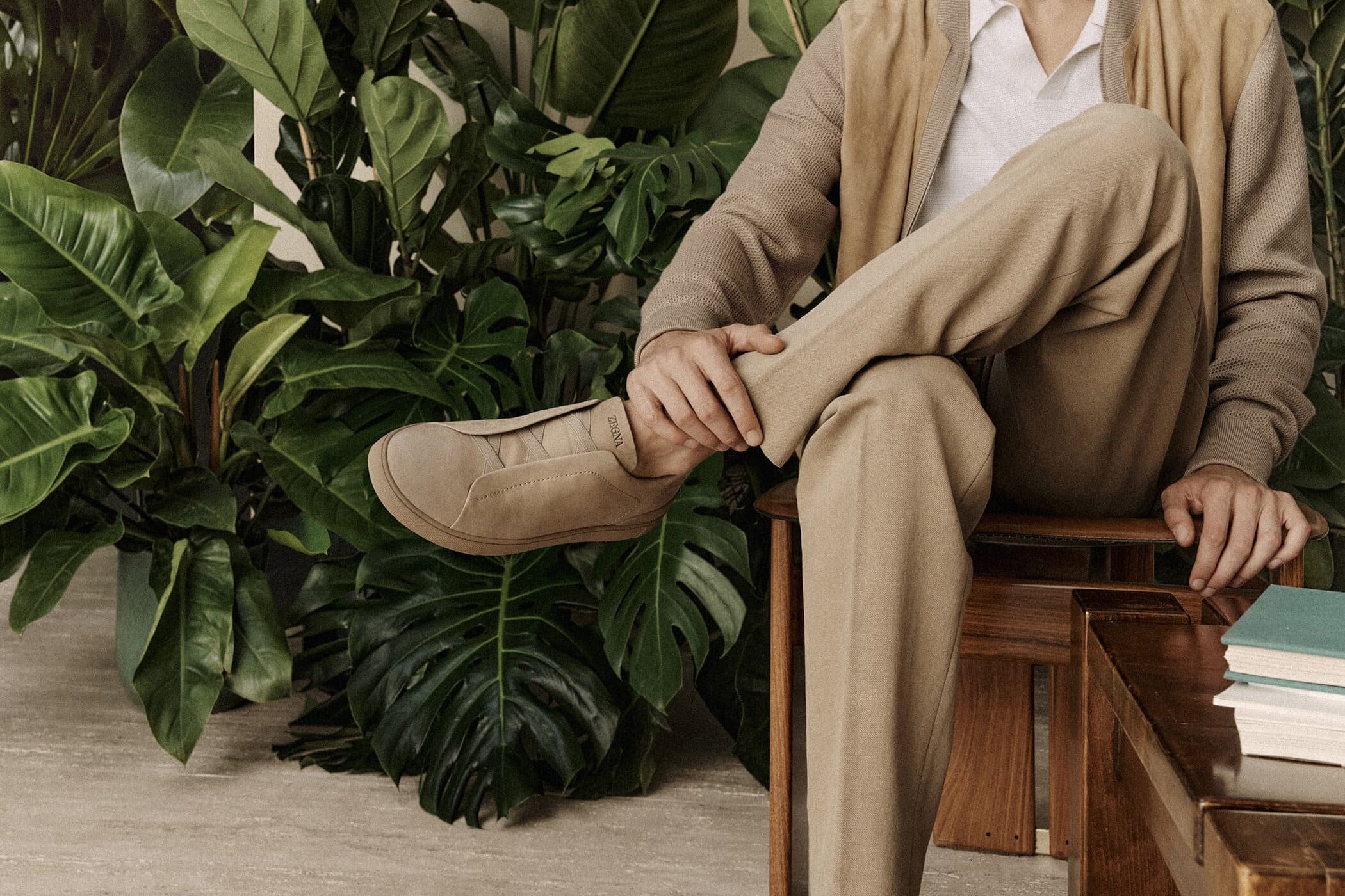 A man in a neutral suit sits with legs crossed, casual shoe visible. Tropical foliage creates a natural, organic background.