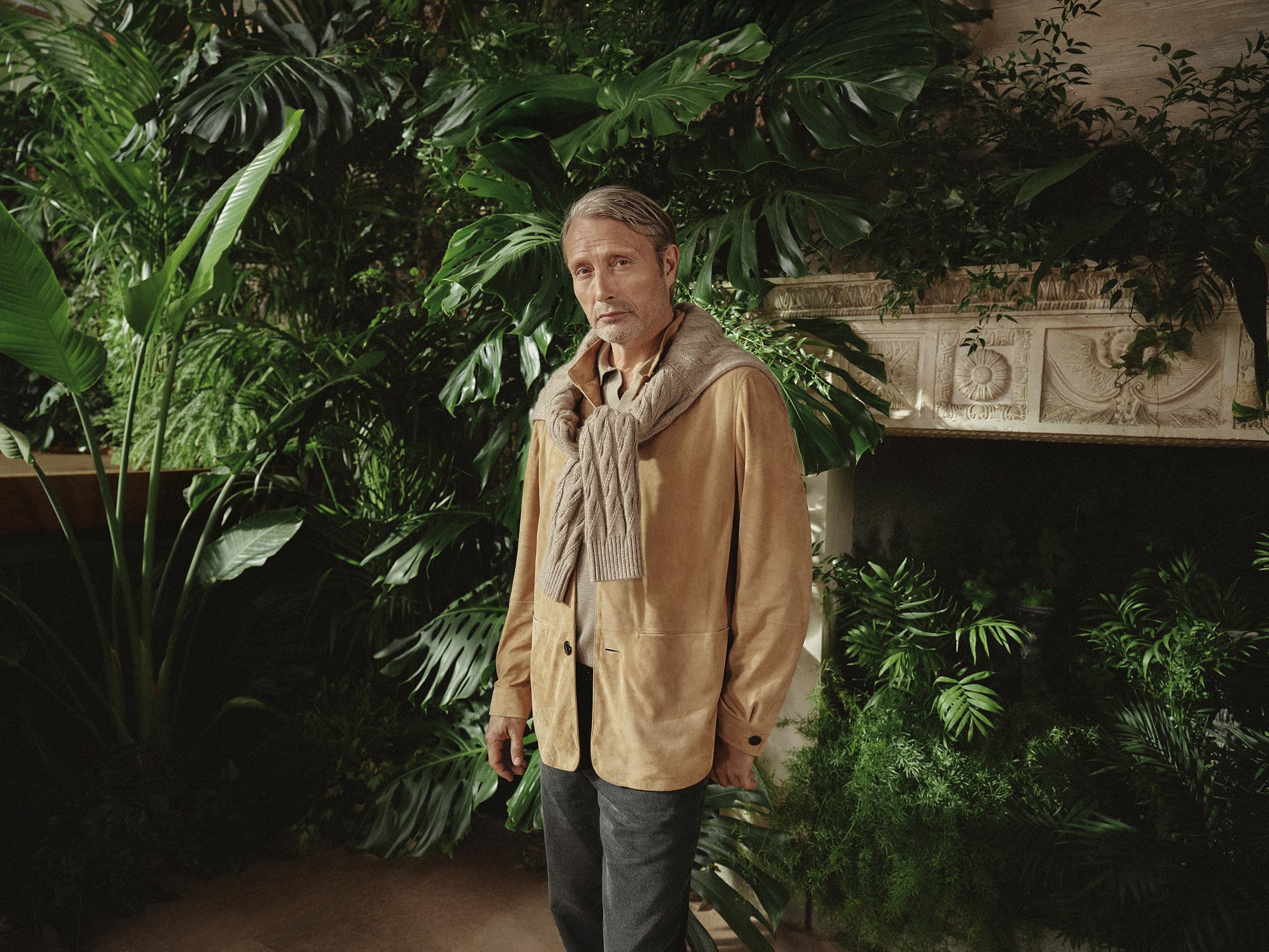 Mature man poses in tan jacket and scarf amid lush green plants and indoor setting, portrait-style composition, natural light. 