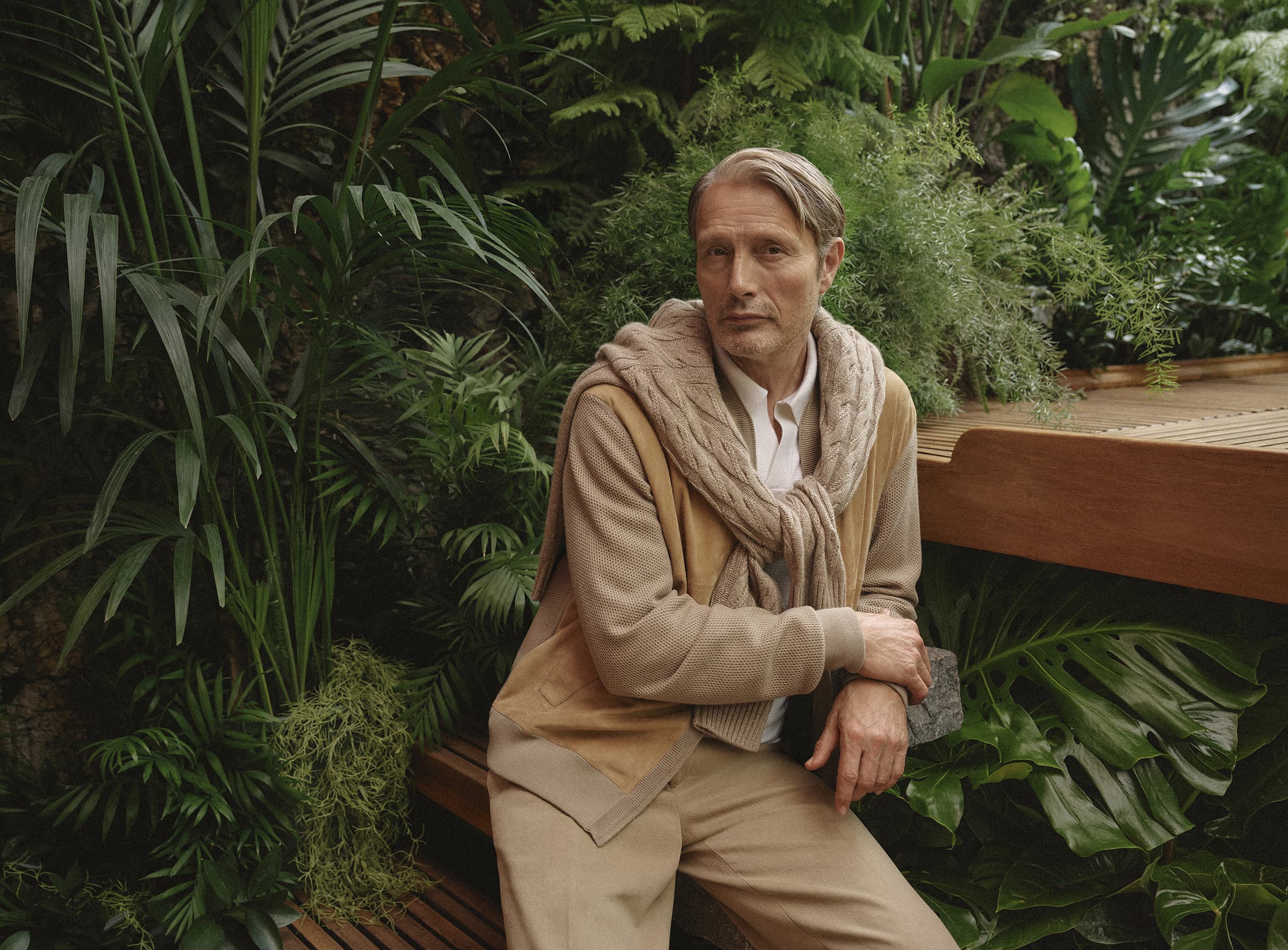 Portrait of a man in a beige cardigan and sweater draped over his shoulders, sitting on a wooden bench amidst lush greenery.