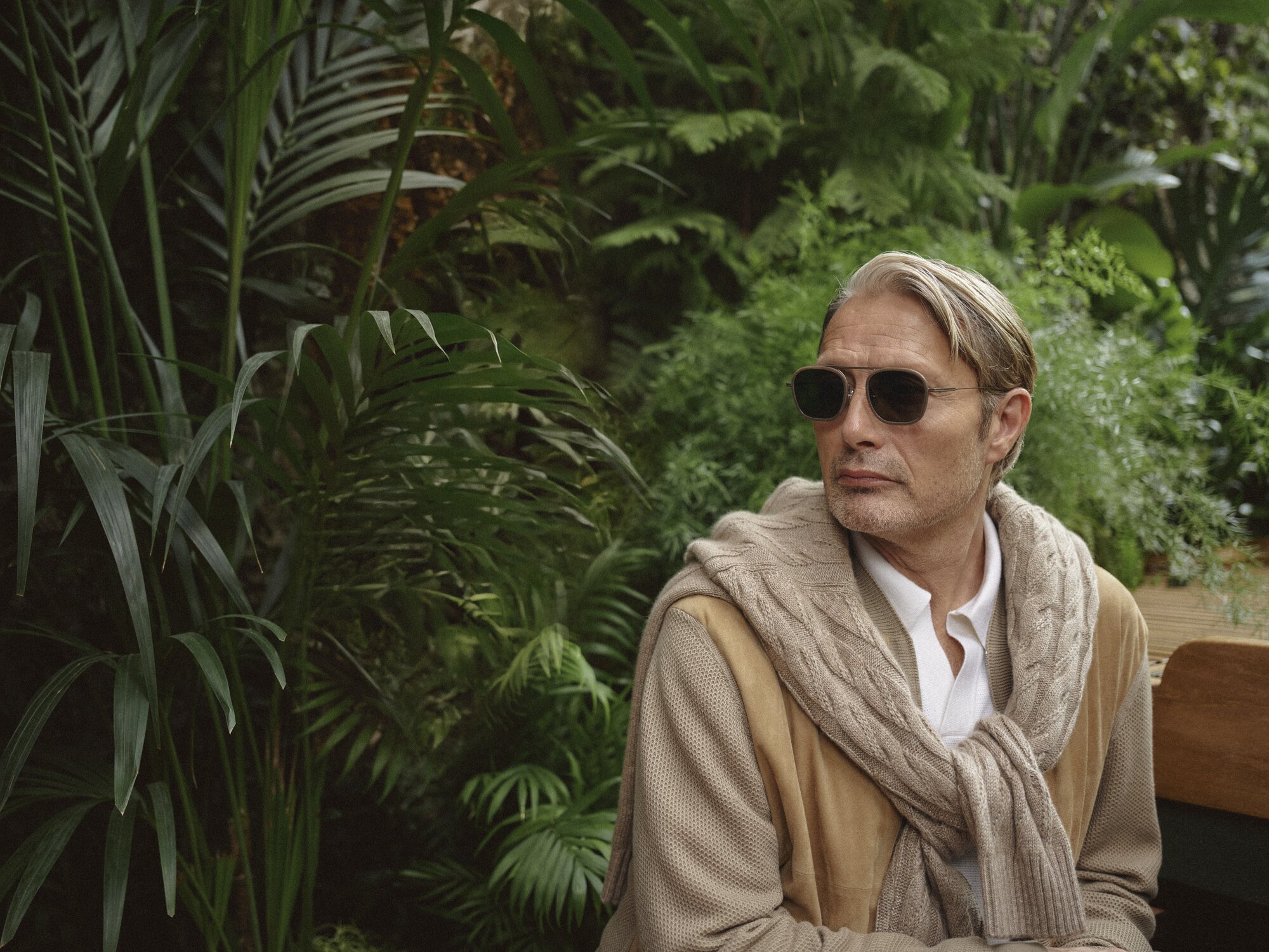 Mature adult man with sunglasses in natural setting. Man wears a cream-colored sweater and tan jacket with lush greenery in the background.