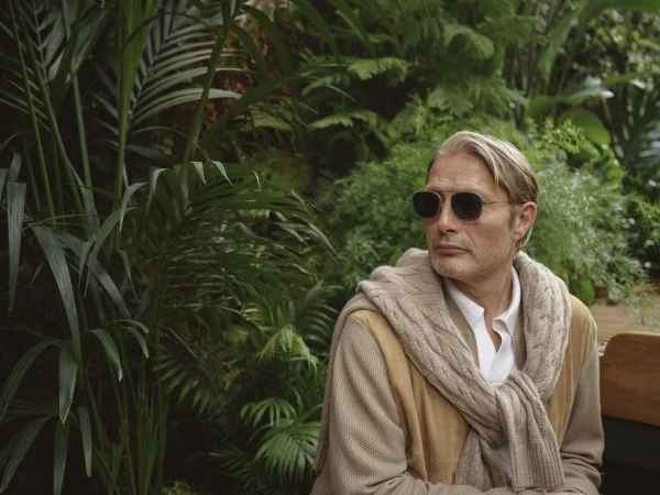 Mature adult man with sunglasses in natural setting. Man wears a cream-colored sweater and tan jacket with lush greenery in the background.