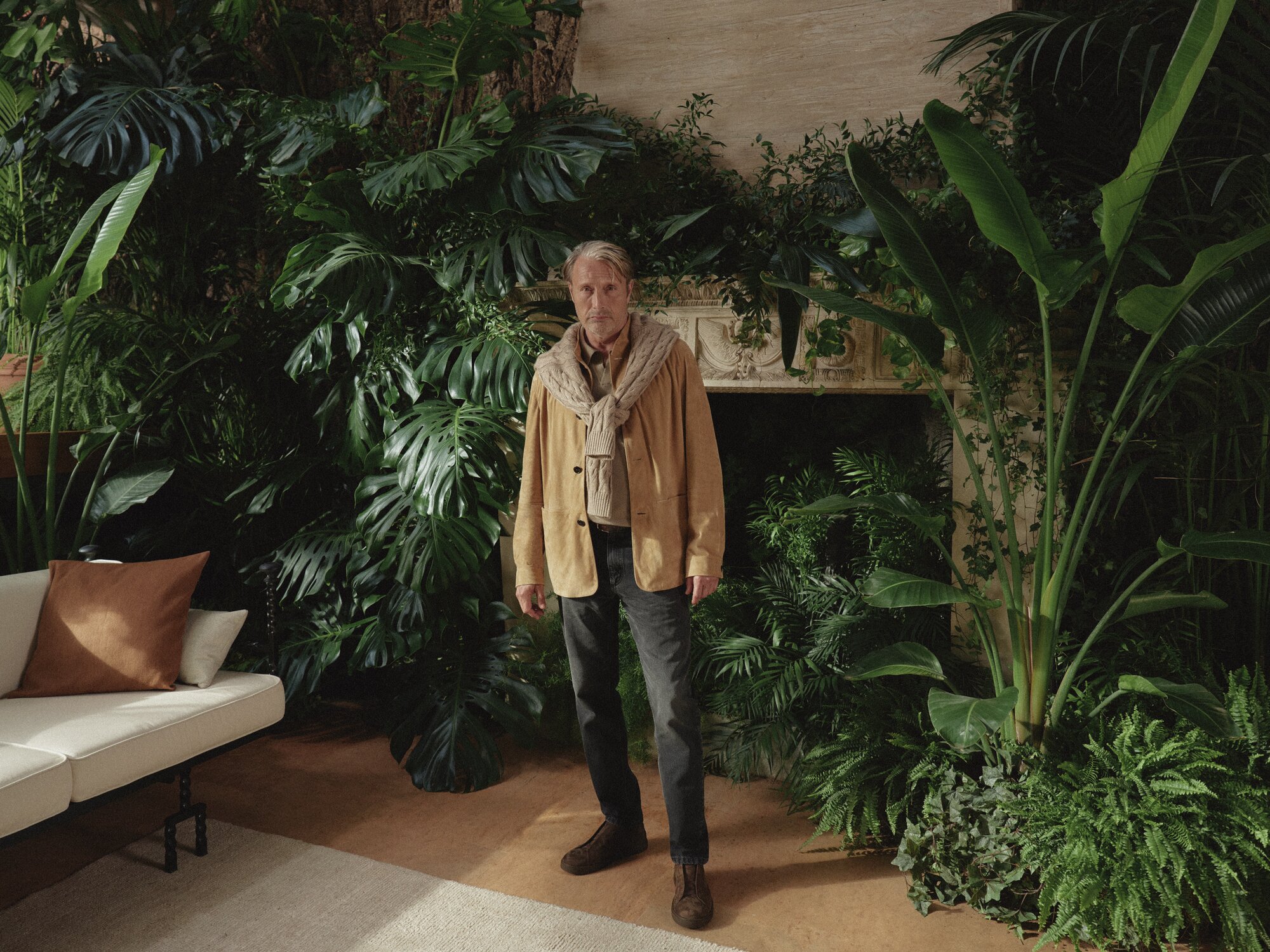 Adult male in a tan jacket and scarf stands centered indoors surrounded by large green plants and natural lighting, with a neutral couch nearby.