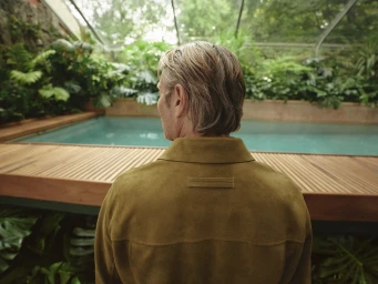 Mads Mikkelsen wearing a tan suede jacket seen from behind overlooking a green indoor pool and lush vegetation.