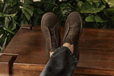 Close-up of brown suede sneakers worn with dark jeans resting on a wooden surface against a lush green leafy background.