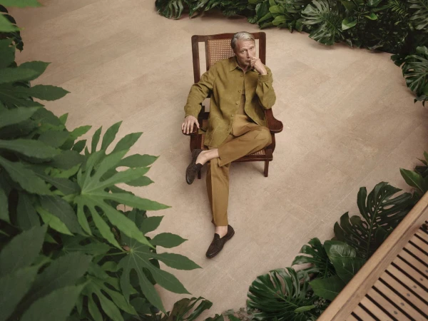 Mads Mikkelsen seated in a wooden armchair wearing an olive green jacket and trousers surrounded by tropical foliage