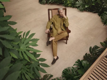 Mads Mikkelsen seated in a wooden armchair wearing an olive green jacket and trousers surrounded by tropical foliage