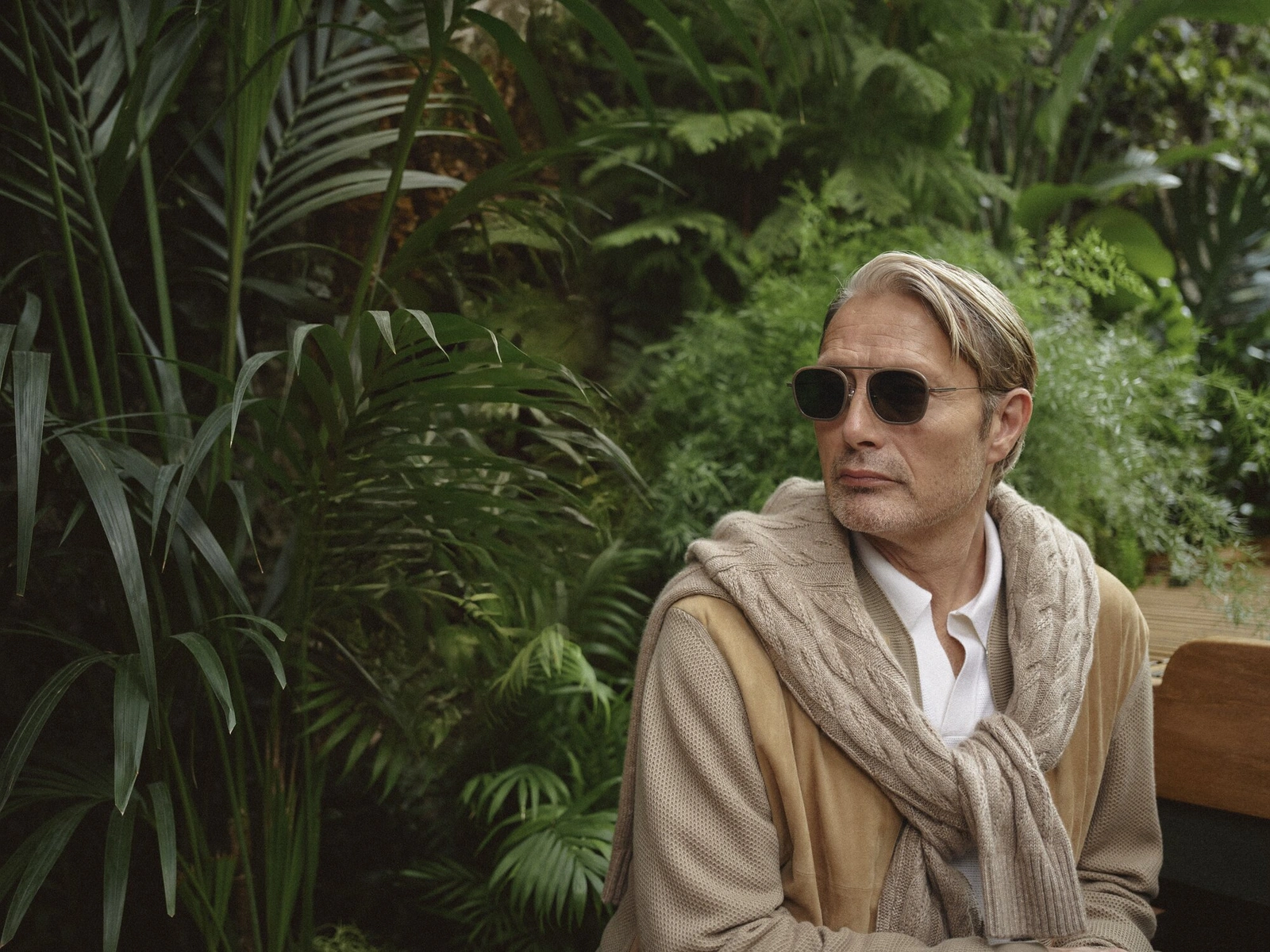 Mads Mikkelsen wearing sunglasses and a cable-knit sweater draped over shoulders amidst lush green foliage for Zegna.