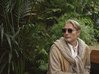 Mads Mikkelsen wearing sunglasses and a cable-knit sweater draped over shoulders amidst lush green foliage for Zegna.