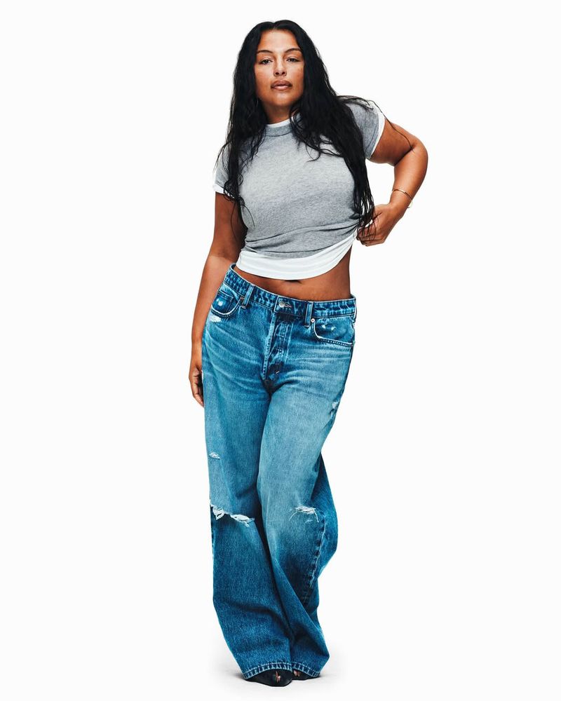 A woman with long dark hair poses in a photo studio wearing a gray t-shirt and ripped, wide-leg blue jeans against a white background.