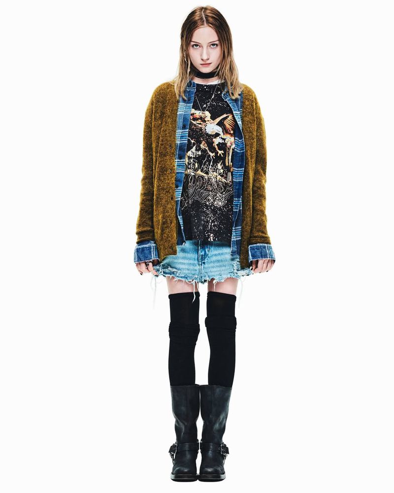 A young woman in a casual fashion outfit poses in a full shot against a white backdrop, wearing a printed tee, skirt, cardigan, and boots.
