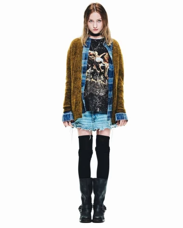A young woman in a casual fashion outfit poses in a full shot against a white backdrop, wearing a printed tee, skirt, cardigan, and boots.