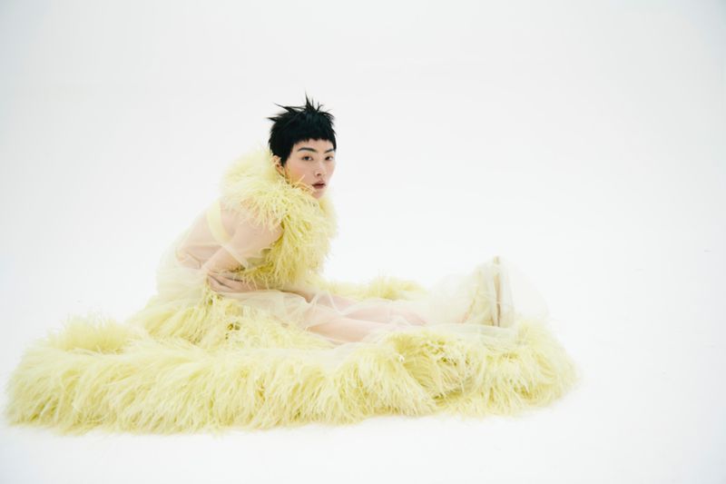 Person in a yellow fur garment poses on a white background, seated with legs outstretched, looking directly at the camera in a studio setting.