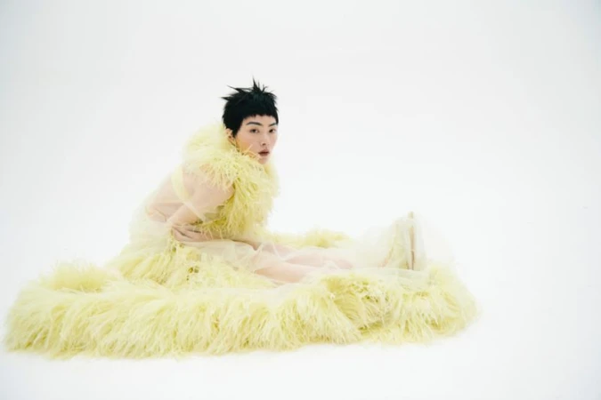 Person in a yellow fur garment poses on a white background, seated with legs outstretched, looking directly at the camera in a studio setting.