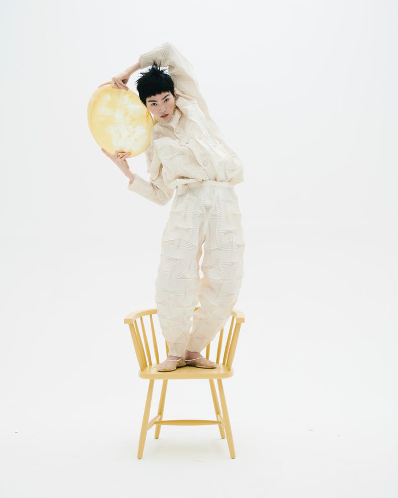 Fashion shot of a young adult female in a textured beige outfit standing on a chair while holding a yellow balloon in a white studio.