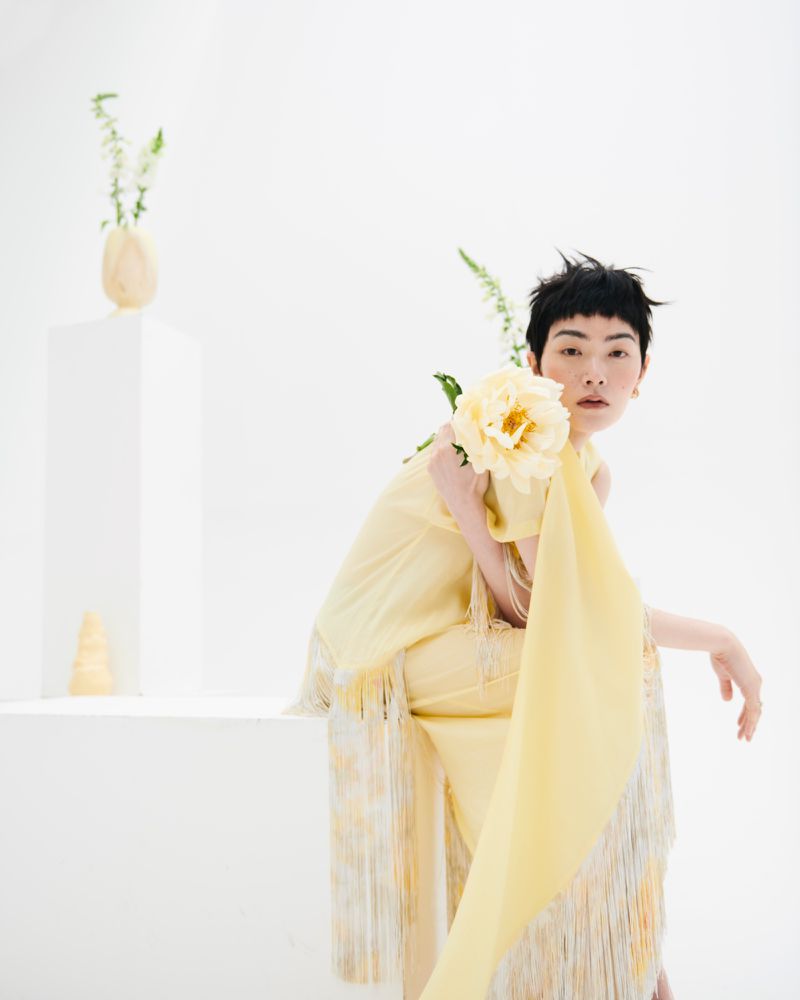 An Asian woman poses in a studio with a yellow flower, wearing a fringed dress against a white background, creating a minimalist fashion portrait.