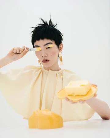 Woman with short black hair poses with yellow-colored eyeshadow and decorative yellow plastic props in a brightly lit photo studio.