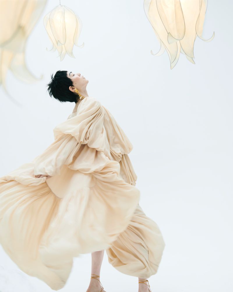 Elegant woman in flowing beige gown poses in a light, bright photo studio with hanging flower-shaped lamps, fashion editorial photography.