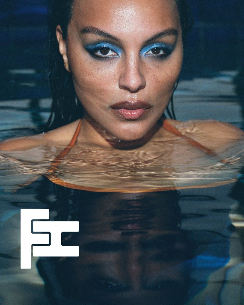 Close-up portrait of a model with dramatic blue eye makeup in a pool of water with a reflection below.