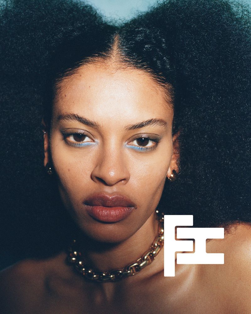 Close-up portrait of a Black woman with an afro hairstyle, wearing a gold chain necklace and blue eyeliner makeup against a dark backdrop.