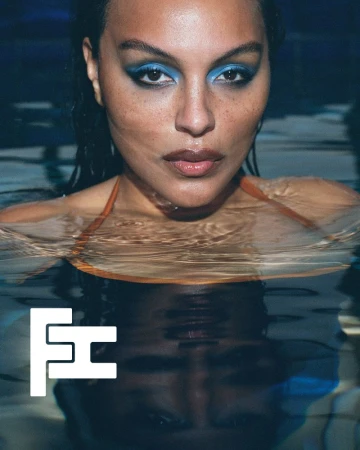 Close-up portrait of a model with dramatic blue eye makeup in a pool of water with a reflection below.