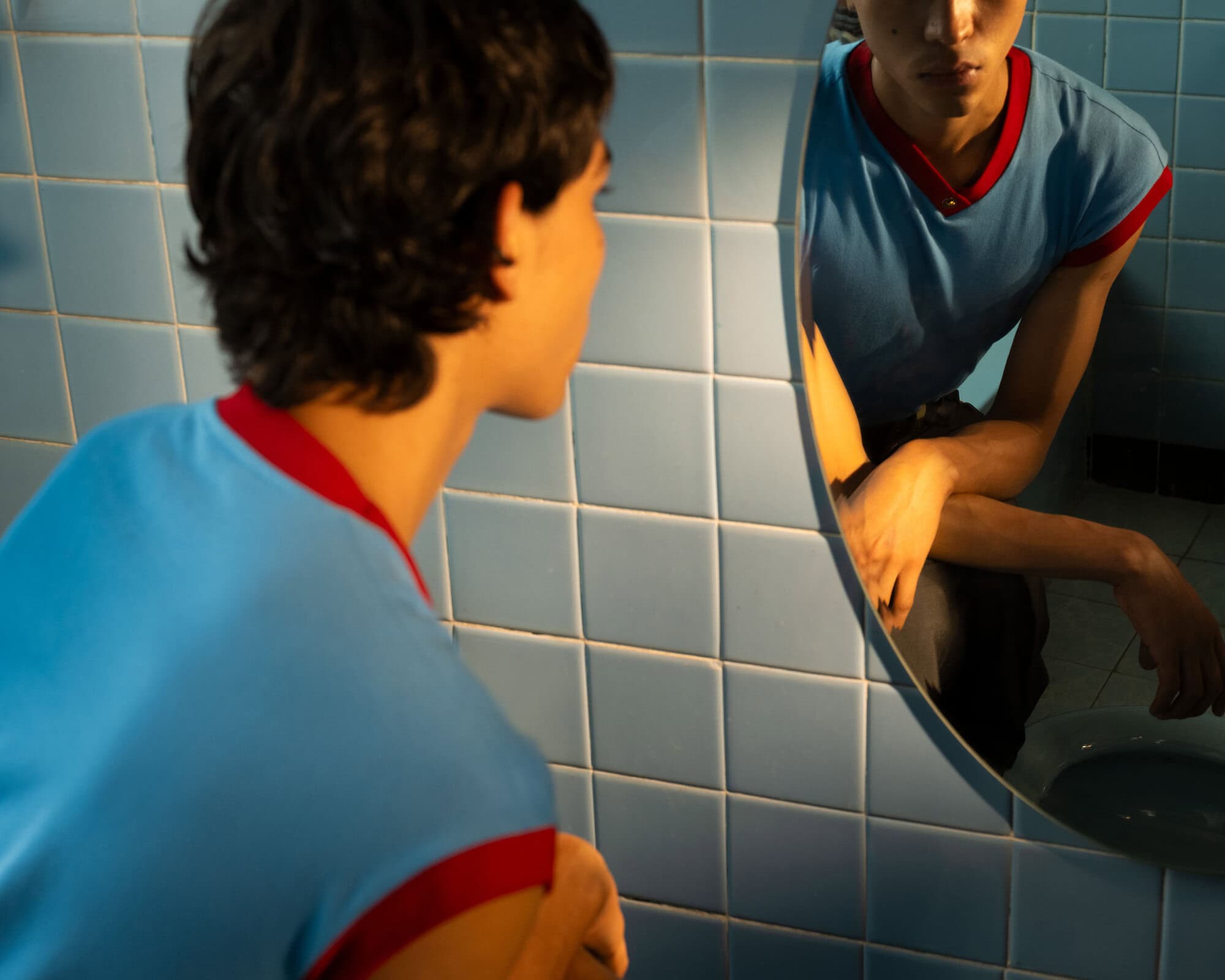 A young adult in a light blue shirt with red trim looks in a circular mirror in a blue-tiled bathroom with warm, golden light.