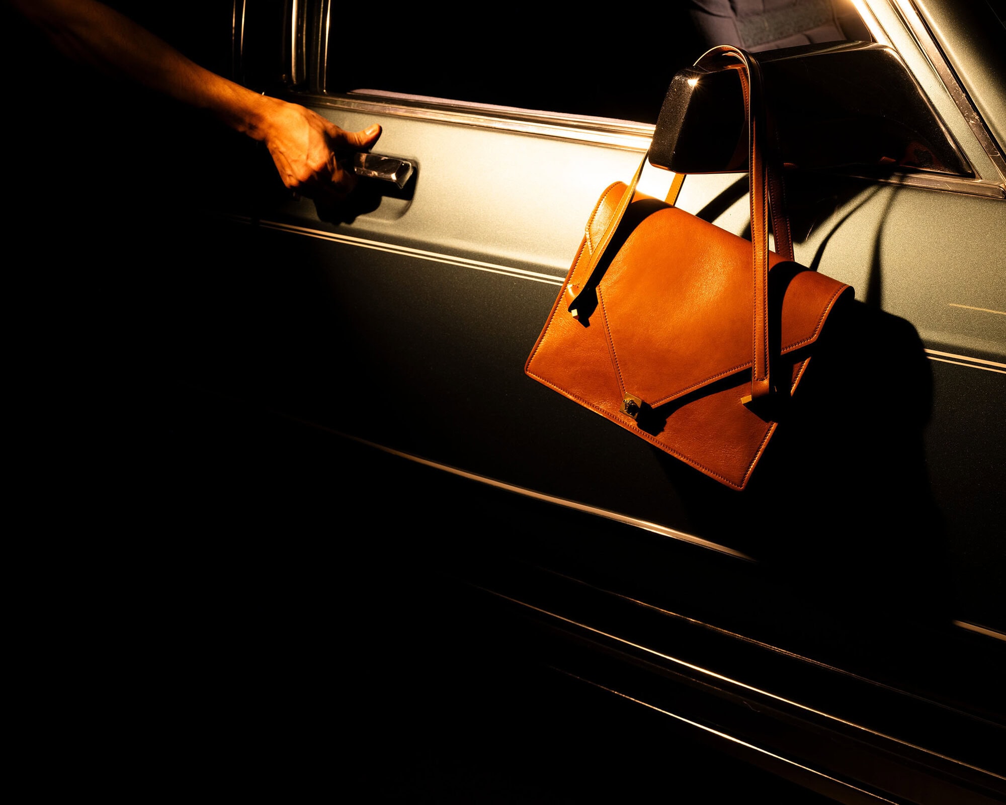 Cropped shot of a woman's arm reaching to open a car door with a brown leather handbag hanging from the car's frame in dark lighting.