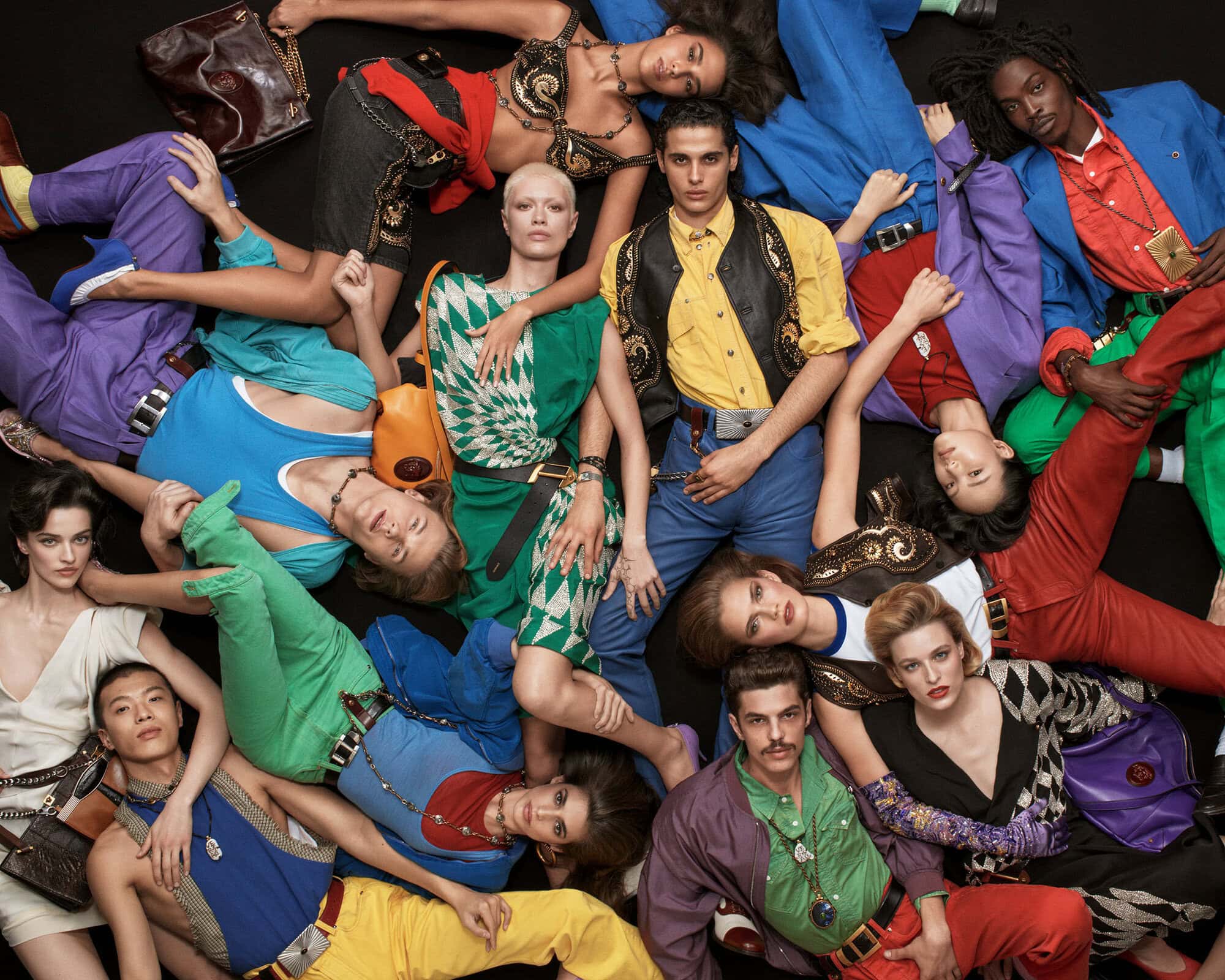 Fashion editorial image featuring a diverse group of models in colorful retro-inspired clothing, posing on a black backdrop with studio lighting.