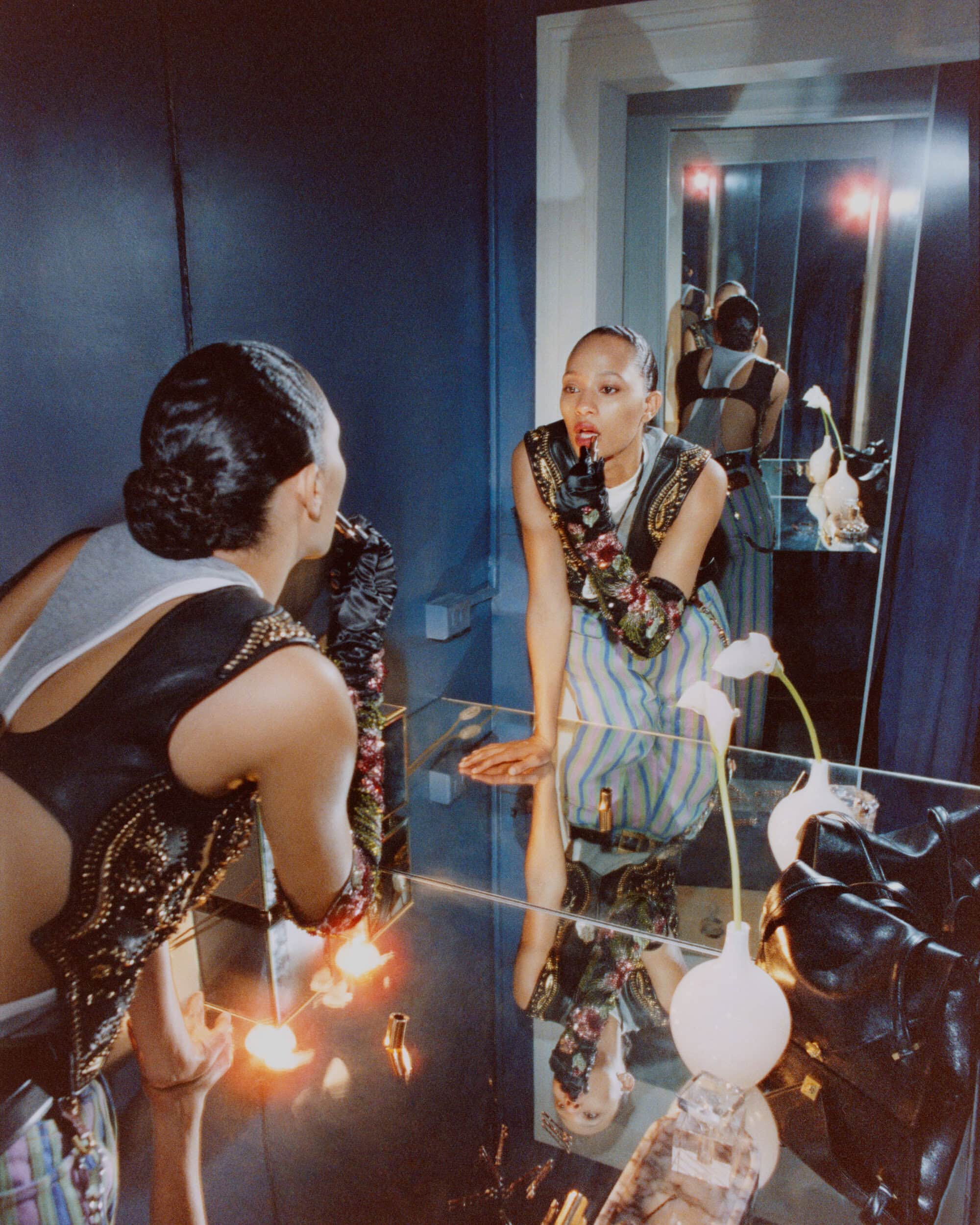 A woman applies makeup in a mirrored room, wearing a fashion-forward outfit, captured in a studio with moody lighting.