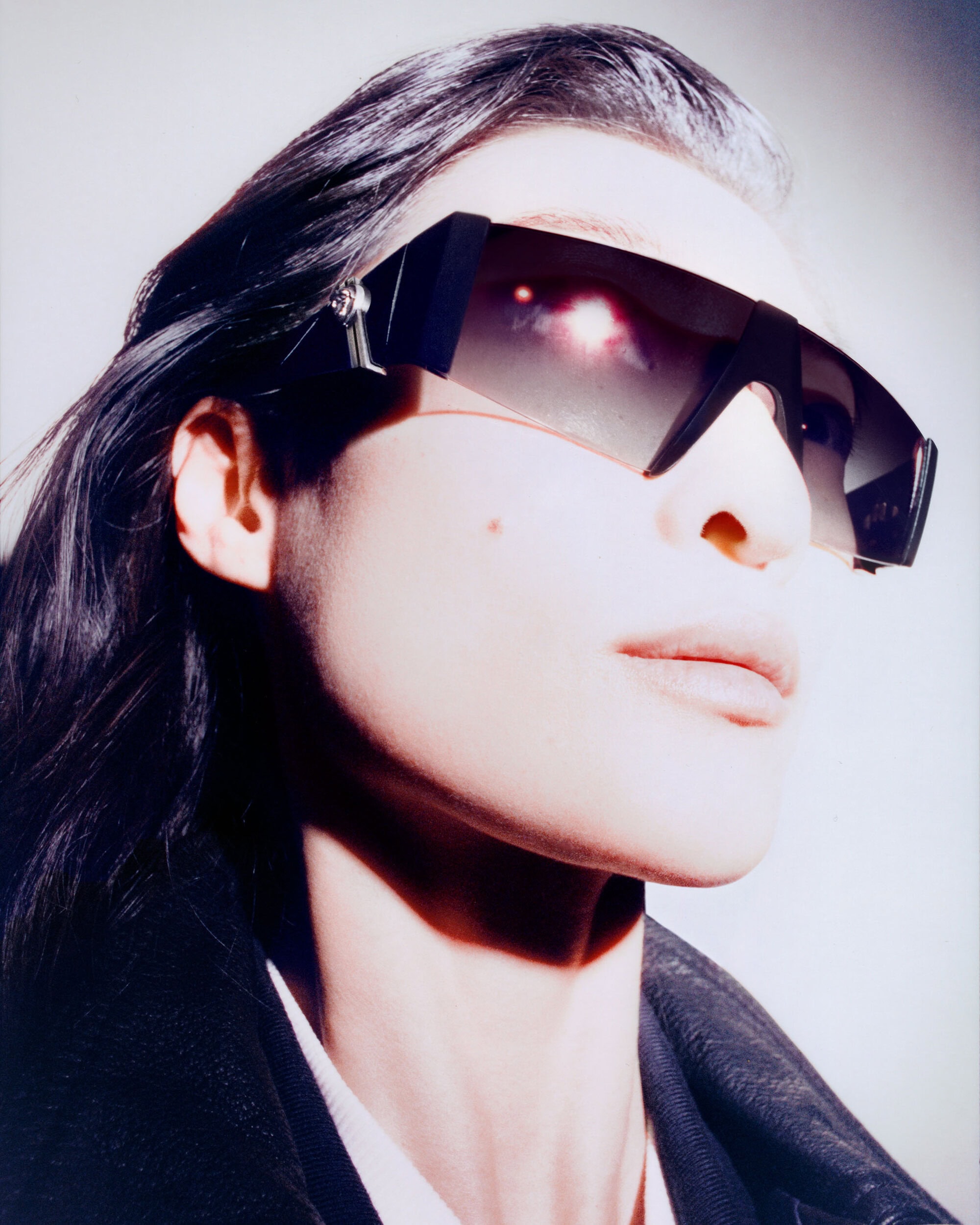 Close-up portrait of a model wearing dark futuristic sunglasses and a black jacket against a plain light background.