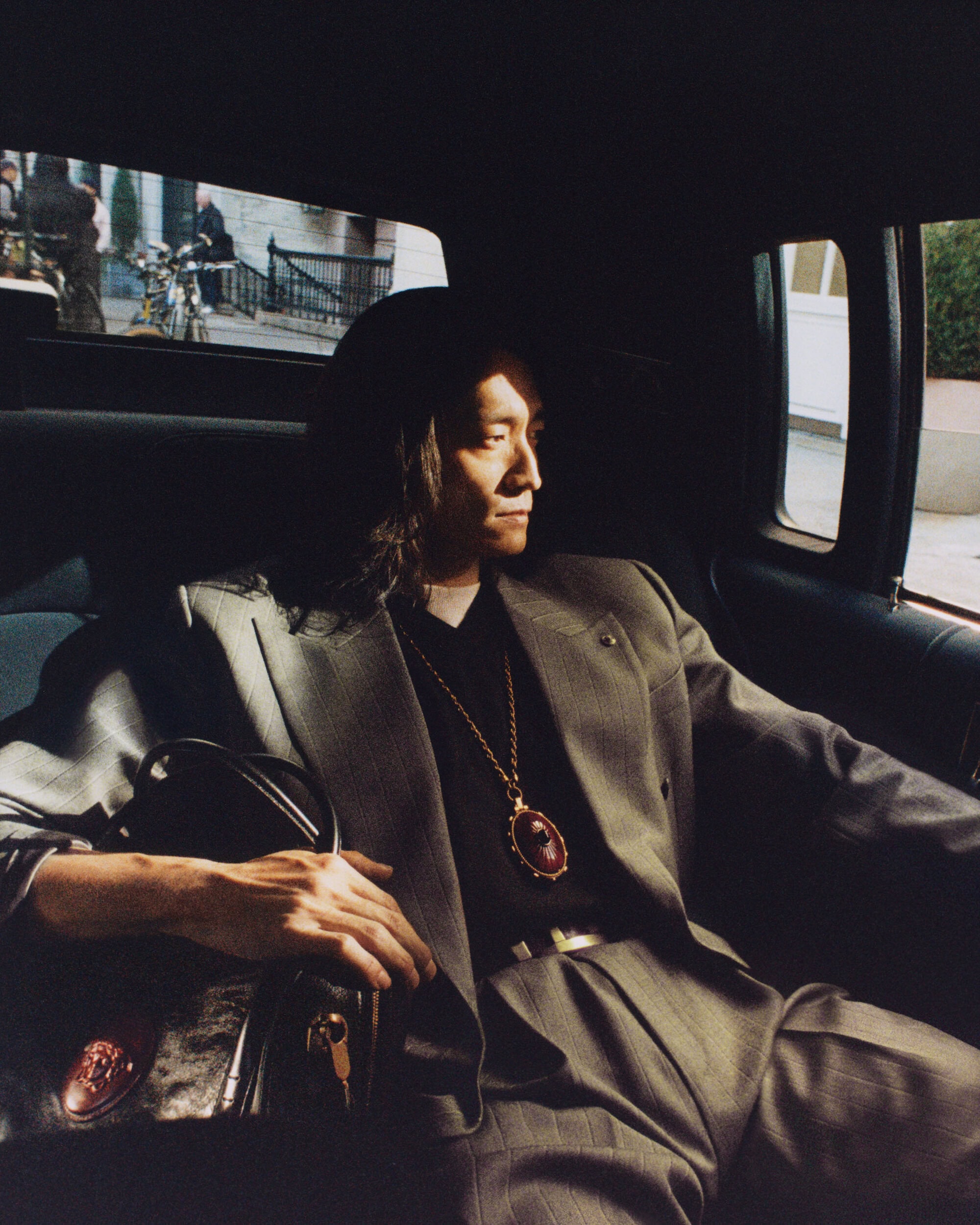 Man in a grey suit sits in the back of a car, looking out the window, holding a purse, with city buildings visible outside.