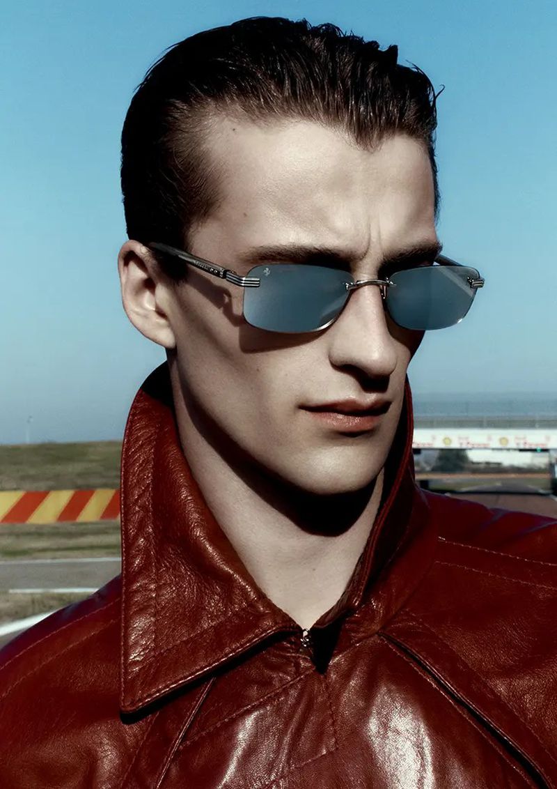 Close-up of a man with dark, slicked-back hair, wearing a red leather jacket and reflective sunglasses outdoors on a racetrack.