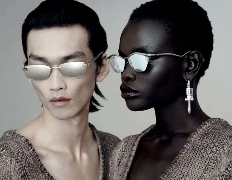 Fashion shot of a dark-skinned woman and light-skinned man wearing sunglasses and draped knitwear in a studio setting with neutral background.