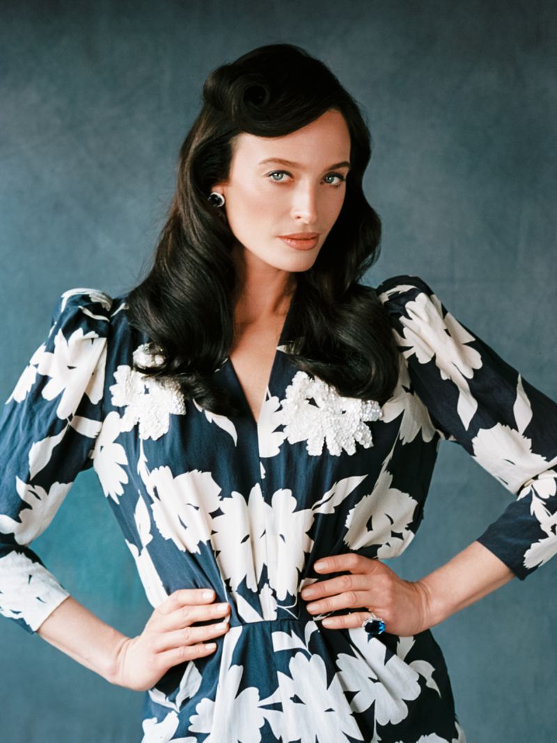 Portrait of a brunette woman in a navy and white floral dress with hands on her hips, styled dark hair, and a blue textured background.