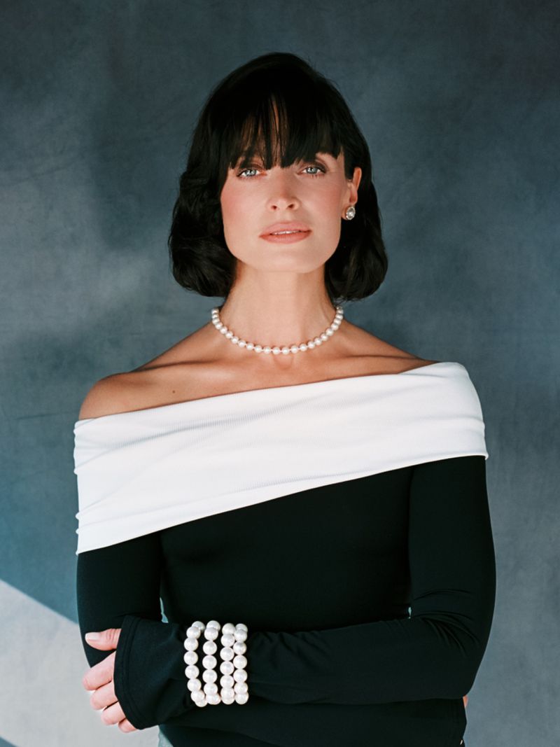Portrait of an adult female with dark hair wearing pearls and a black and white off-the-shoulder dress against a grey backdrop.