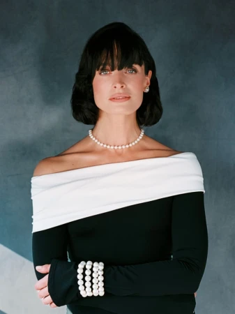 Portrait of an adult female with dark hair wearing pearls and a black and white off-the-shoulder dress against a grey backdrop.
