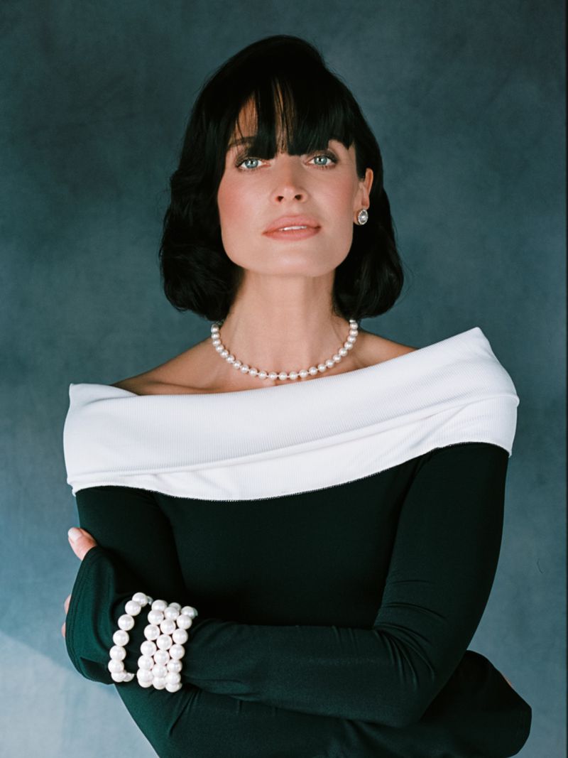 Elegant woman with short black hair, wearing a black and white off-the-shoulder dress and pearl jewelry, posing in a studio.