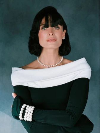 Elegant woman with short black hair, wearing a black and white off-the-shoulder dress and pearl jewelry, posing in a studio.