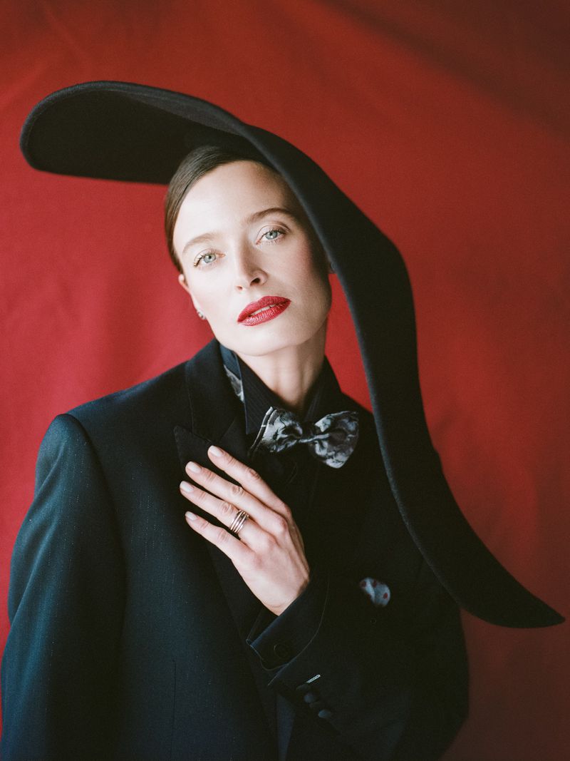 Elegant woman in a black blazer, patterned bow tie, and a dramatic black hat, set against a red backdrop in a fashion portrait.
