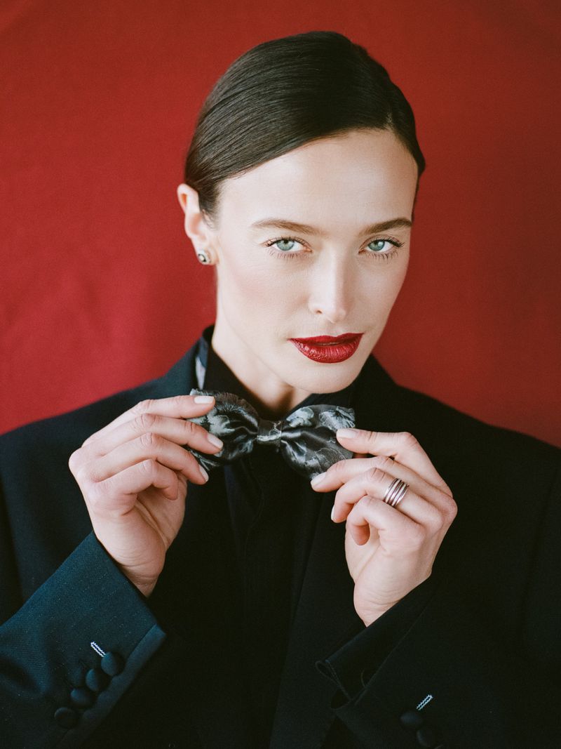 Portrait of a stylish woman with dark hair and bright red lipstick adjusting a grey bow tie while wearing a black blazer on a red background.