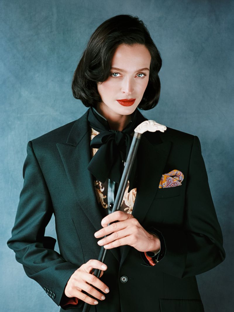 Portrait of a stylish woman in a dark suit, holding a cane, with vibrant red lipstick against a textured blue backdrop.