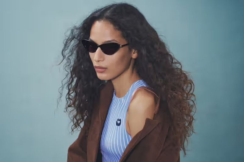 Model with long dark curly hair wearing sunglasses and a brown jacket over striped top against a plain studio background.
