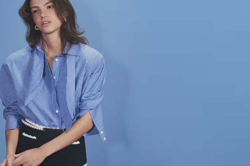 Brunette woman in blue striped shirt and black skirt posing against light blue background in a fashion shot with medium framing.