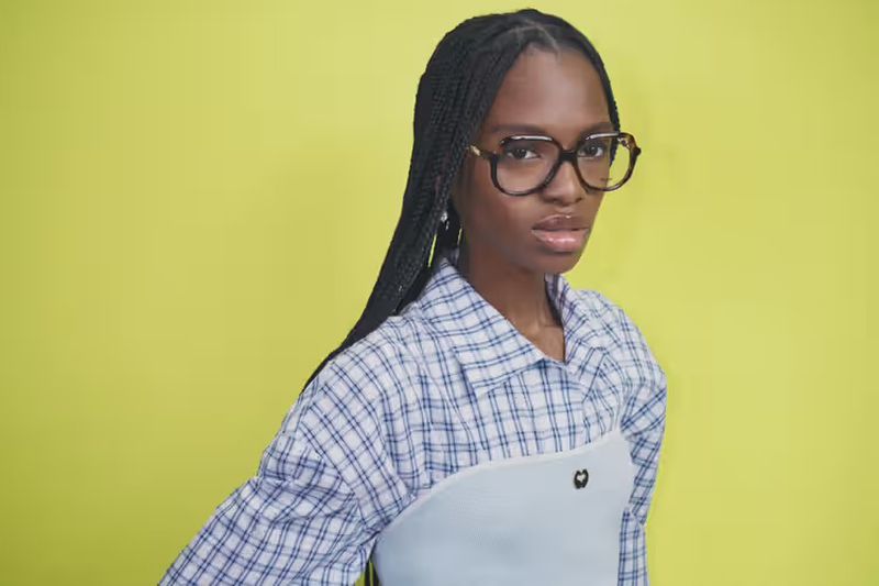 Portrait of a Black woman with braided hair, wearing glasses and a blue plaid shirt, posing against a bright yellow background.