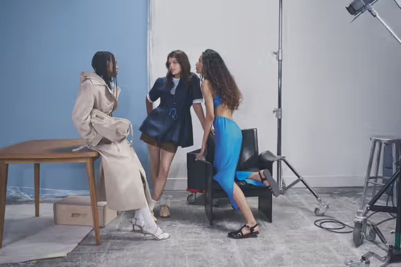 Three diverse adult females in stylish clothing interact on a photo studio set with lighting equipment, a table, and blue and white backdrop.
