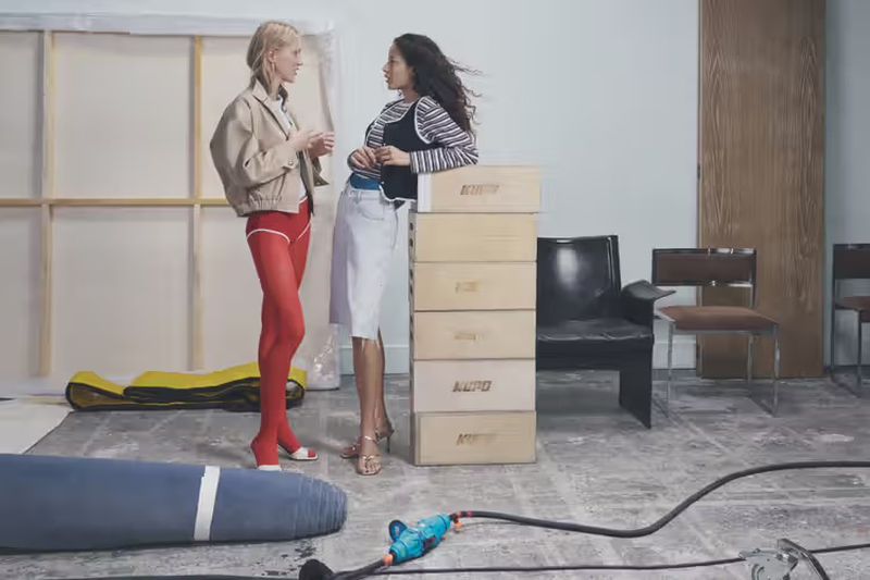 Two women in stylish outfits chat indoors against a backdrop of stacked boxes, studio props, and minimalist furniture in a casual setting.