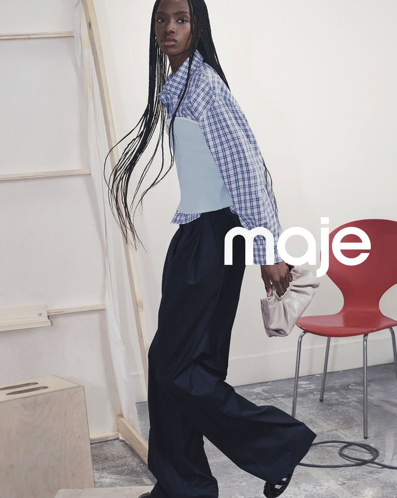 Fashion shot of a Black woman in a plaid top and navy pants, holding a purse, with a red chair and wooden accents in the background.