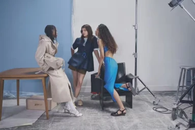 Three models in a photo studio posing near a wooden table and black chair with studio lighting equipment