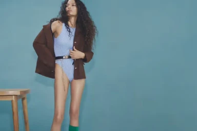 Model with long dark curly hair wearing a blue striped bodysuit and a brown jacket against a solid teal background