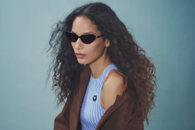 Model with long dark curly hair wearing sunglasses and a brown jacket over striped top against a plain studio background.